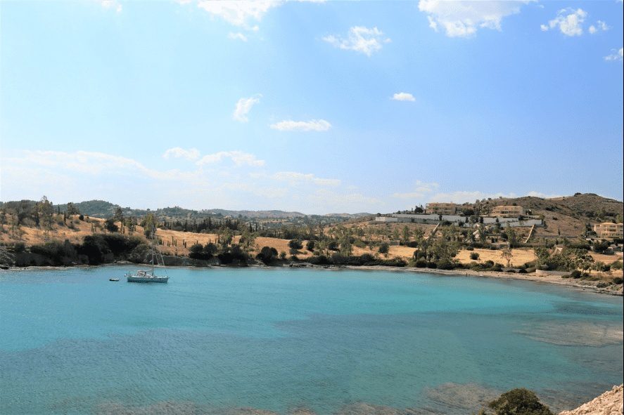 A scenic coastal bay with clear turquoise waters, rolling dry hills in the background, and a lone sailboat anchored near the shore.