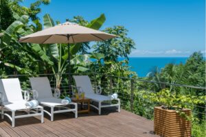 White sun loungers and a beige parasol on a wooden deck overlooking a lush green tropical forest and the sea.