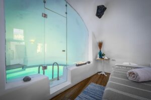 Luxury spa and wellness room featuring a massage bed with rolled towels and a glass partition looking into an indoor pool with lit candles.
