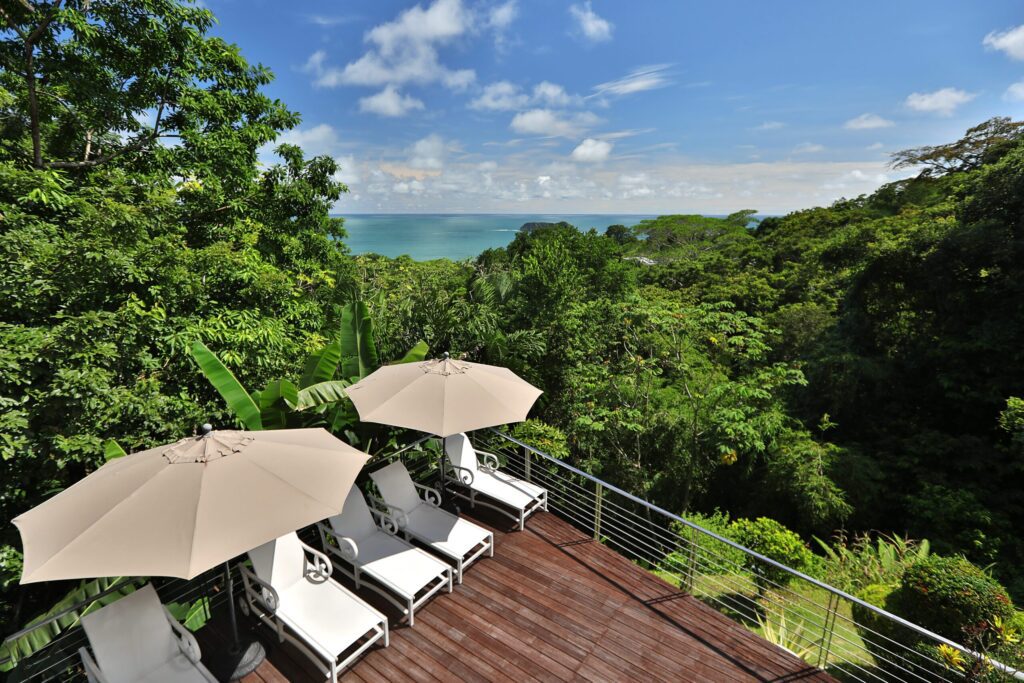 Wooden balcony deck with white lounge chairs and umbrellas overlooking a lush green rainforest canopy and ocean view.