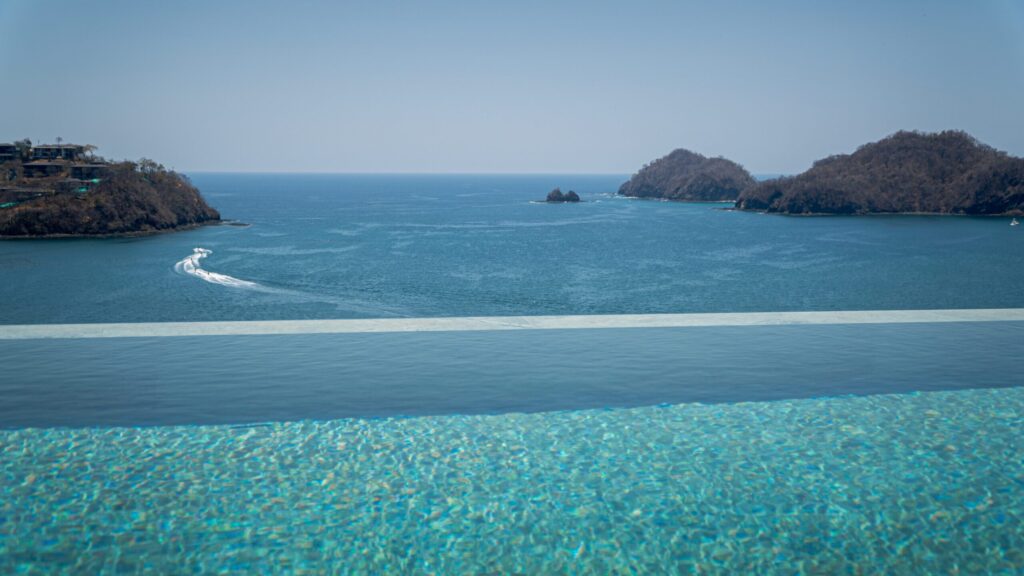 Edge of a luxury infinity pool overlooking a calm blue ocean with distant coastal islands and a boat wake.