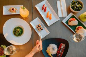 Overhead flat-lay view of gourmet sushi rolls, nigiri, bao buns, seaweed salad, and cocktails spread across a dining table.