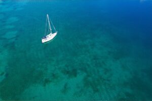 High-angle aerial view of a white sailboat anchored in calm, clear turquoise and blue sea water.