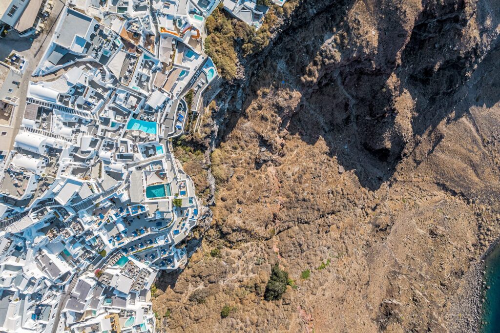 Overhead aerial drone view of white Cycladic architecture and blue pools built along the rugged cliffside terrain in Santorini.