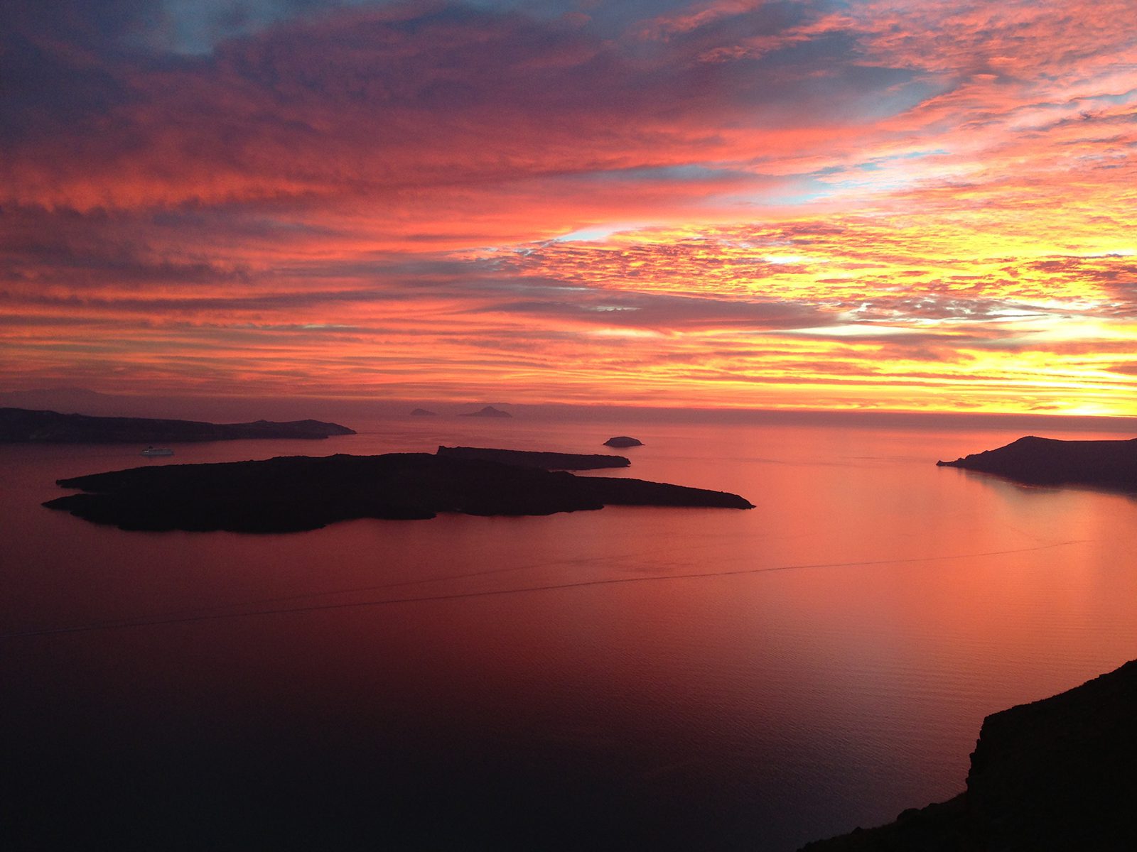 Breathtaking panoramic sunset view over the Santorini caldera with vibrant orange skies reflecting on the sea and dark volcanic islands in the background.